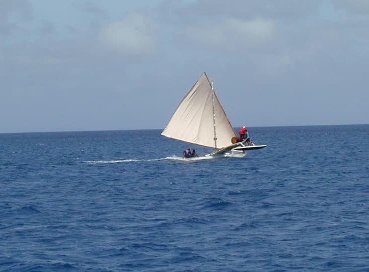 A carved wooden sailing canoe with a colourful sail crossing calm water between two Bijagós islands