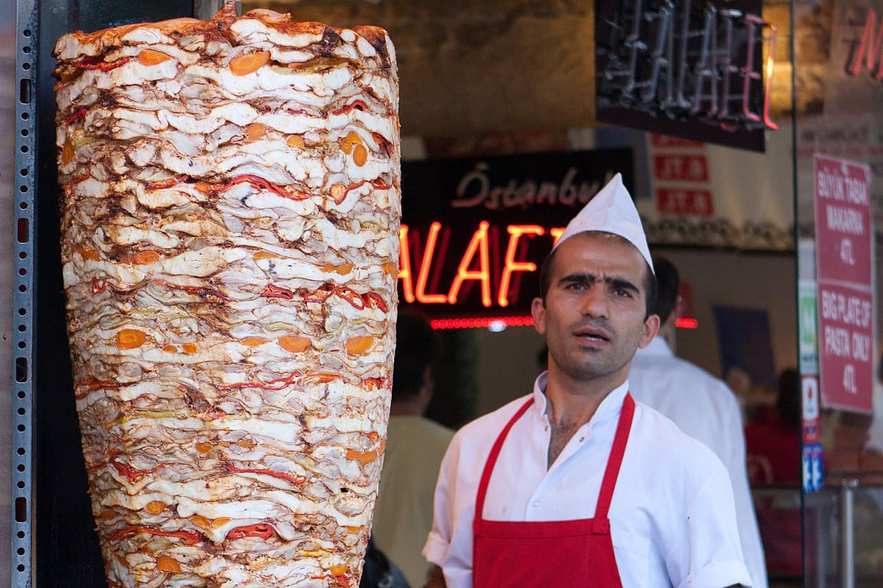 A tall vertical doner kebab spinning slowly in front of a flame in a Turkish kitchen